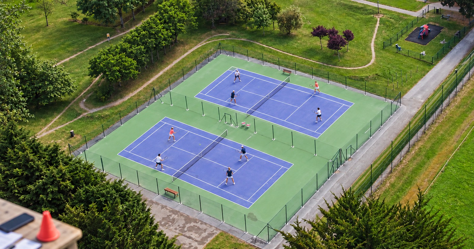 Vue en hauteur de deux matchs de tennis amateur sur des courts voisins, près d’une table d’organisation.